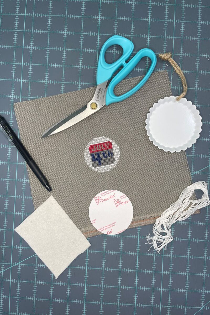 Supplies to finish a round cross stitch piece. This includes, scissors, a frixion pen, round sticky board, batting, and embroidery floss.