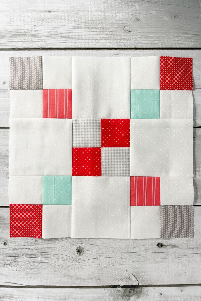 A modified 9 patch quilt block with four patches in the corners and center to make two diagonal lines of pink, red, teal, and grey on a white background fabric.