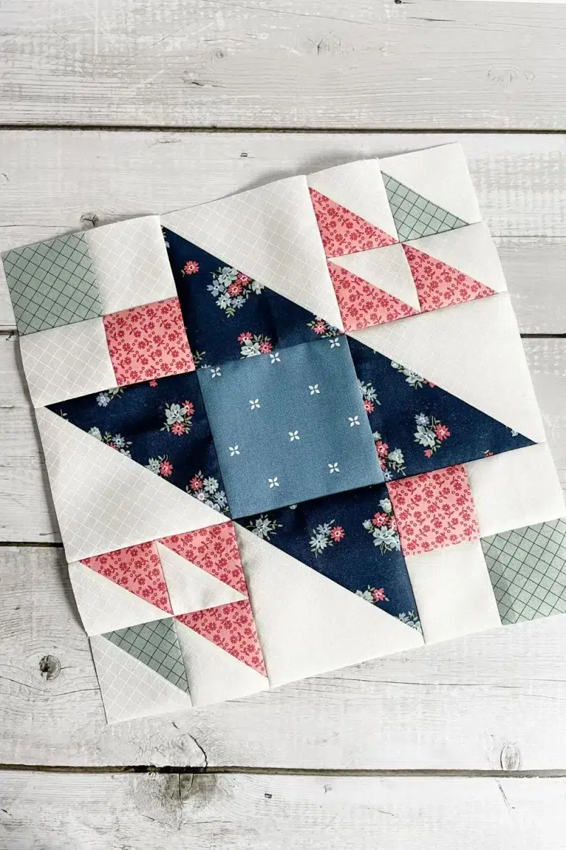 A 12 inch quilt block designed by Barbara Groves. The block is the Orchid block from month 7 of Designer Mystery. It is shown in white, pink, navy, and sage green fabrics.