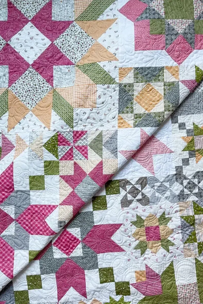 Folded quilt showing many of the star blocks from the Barn Star Sampler quilt in pink, grey, green, and yellow