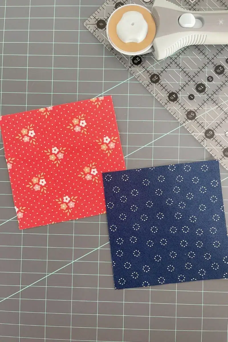Two fabric squares, a rotary cutter, and a quilting ruler used to make Hourglass quilt block units.