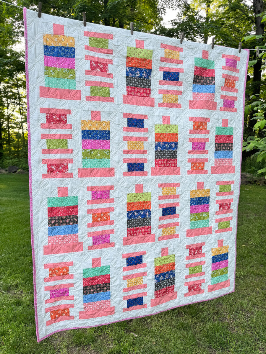 Side view of Strippy Spools jelly roll quilt made with Cali & Co fabric and coral Bella Solid spools, hanging outdoors in natural light.