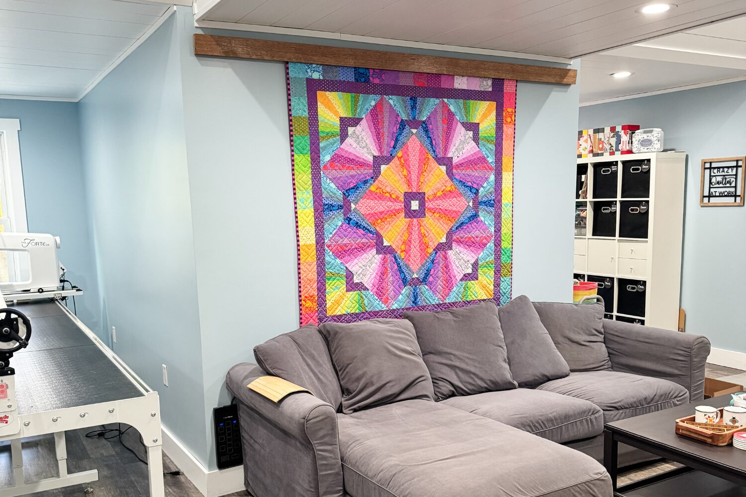 Couch with a quilt hanging above it and a small table holding cross-stitch supplies.