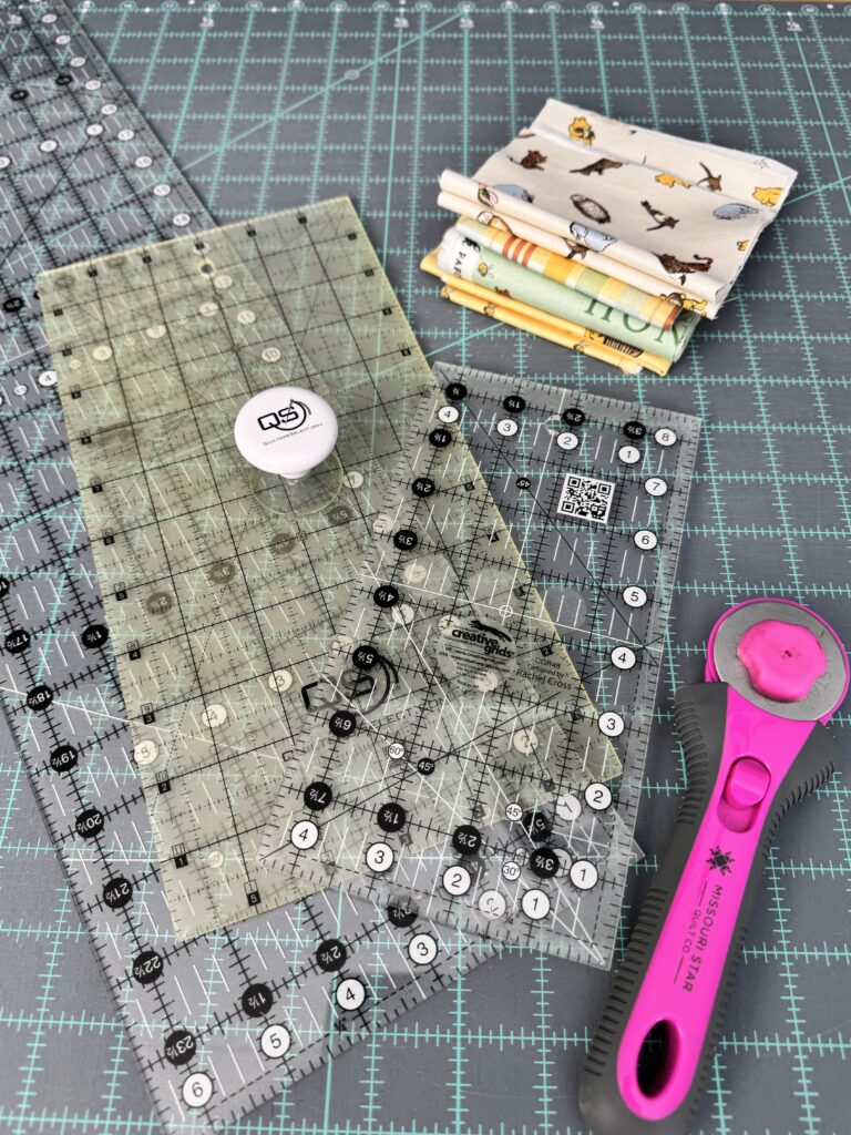 Three quilting rulers fanned on on a Creative Grids cutting mat with a rotary cutter and a neatly stacked pile of fabric