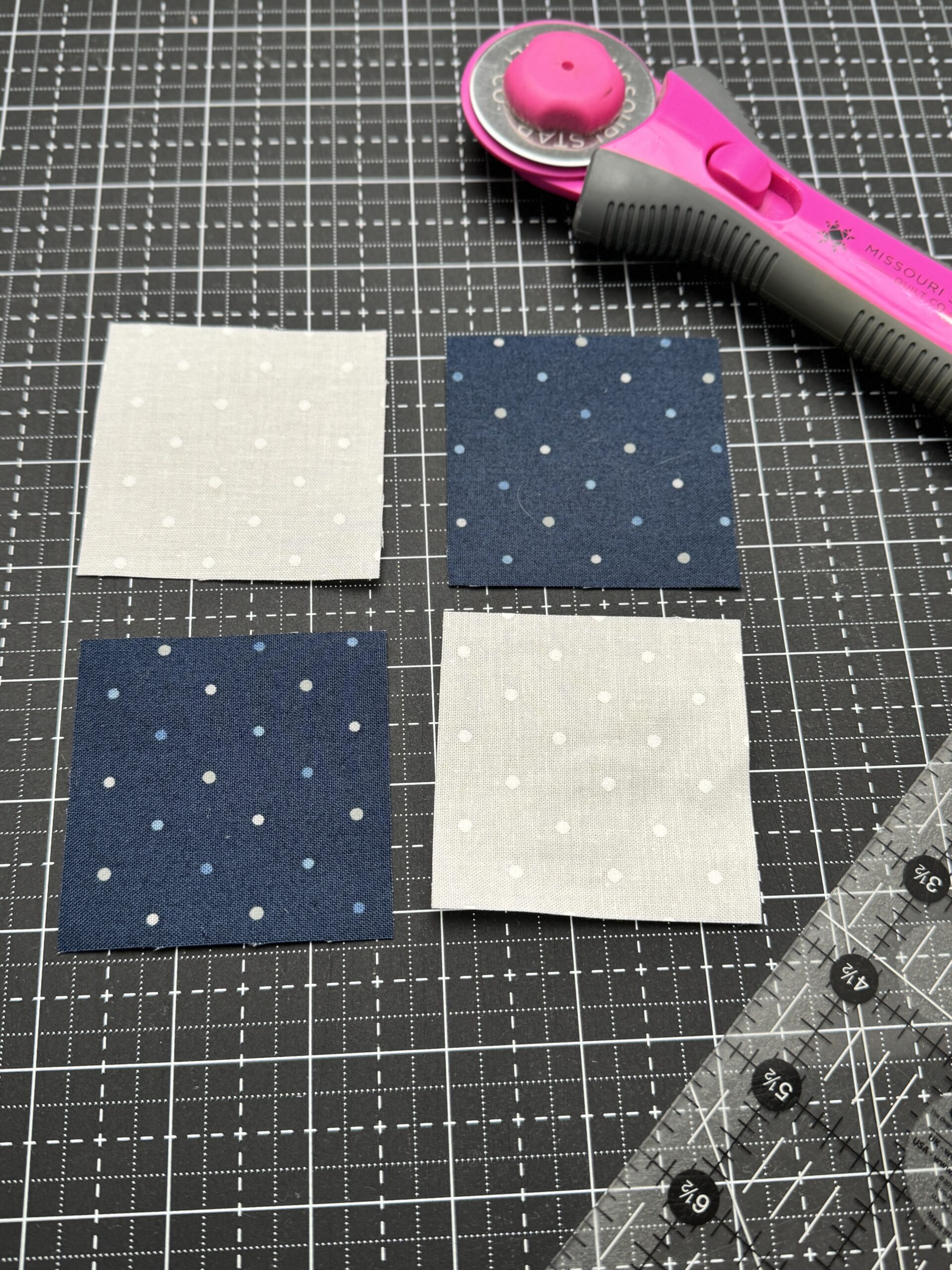 Two white and two blue fabric squares arranged on a black cutting mat with a rotary cutter and quilting ruler nearby