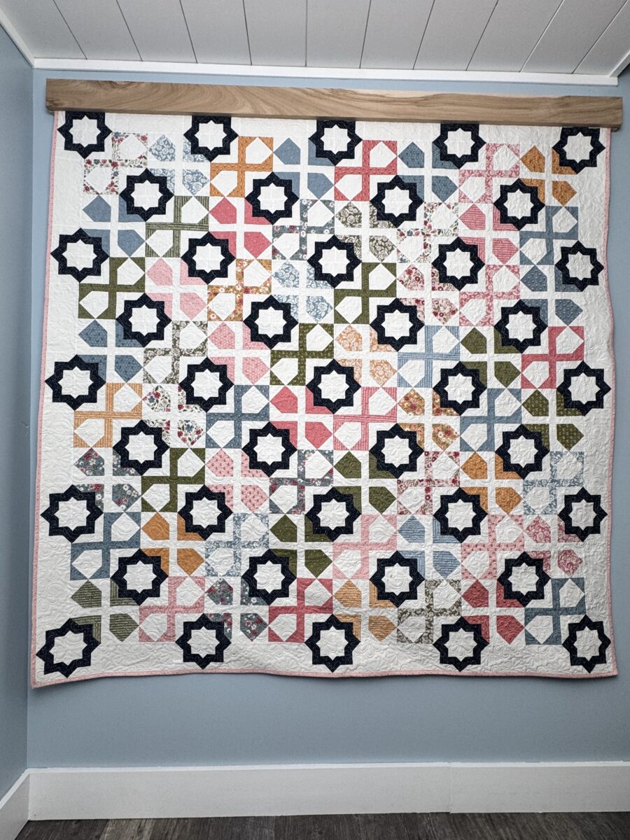 Full view of the Dreamer quilt hanging on a wall showing geometric blocks and navy star accents in Wild Honey fabrics.
