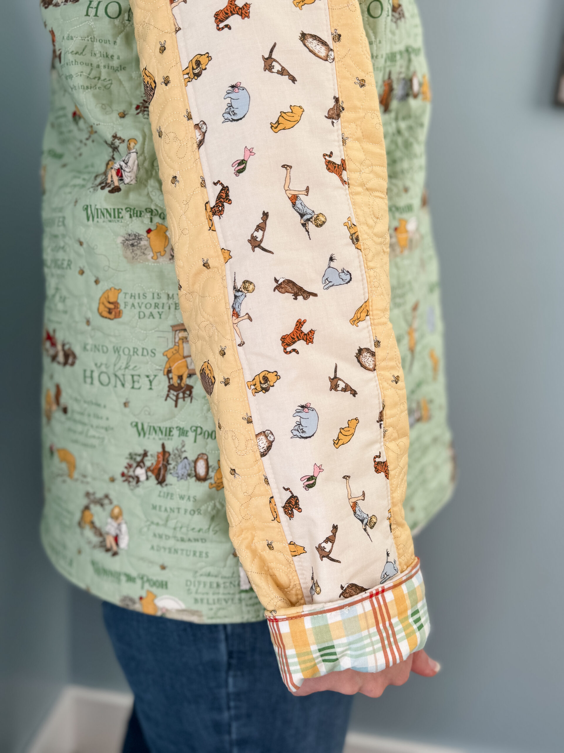 Close up of quilt coat sleeve with cuff folded back showing bright plaid lining and tossed Winnie the Pooh character accent strip.