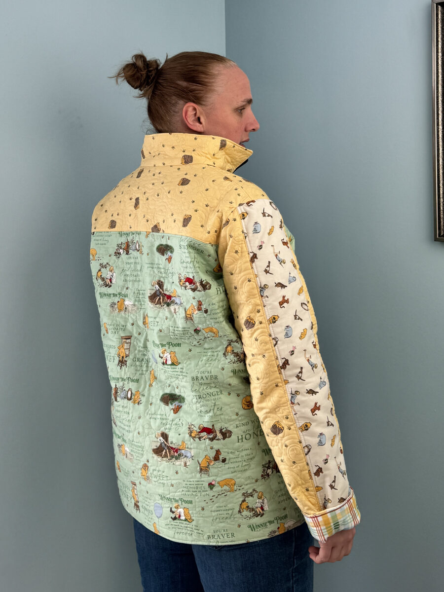 Quilter wearing a Winnie the Pooh themed Bella quilt coat, shown from the back and side to display the green main print and overall silhouette.