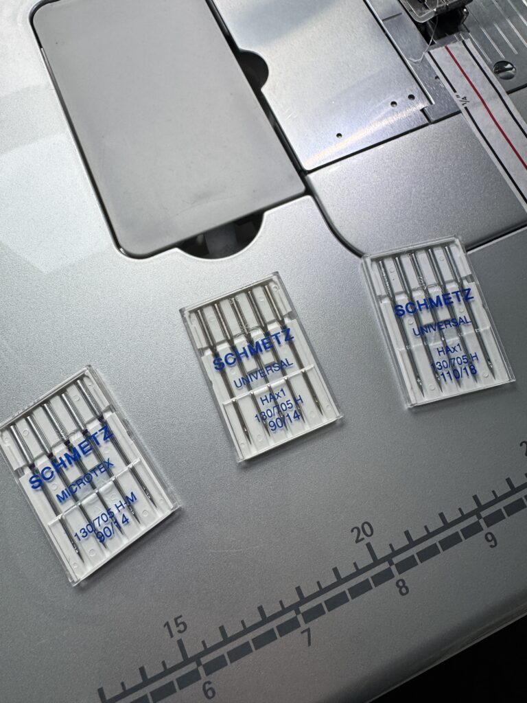 Three Schmetz needle cases arranged on a sewing machine bed showing different needle types for quilting