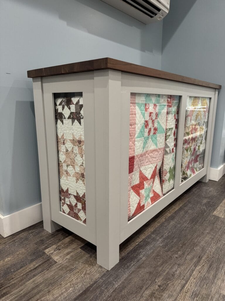 Side view of the DIY quilt cabinet build showcasing a quilt through the glass on the end and multiple quilts on the front glass display.